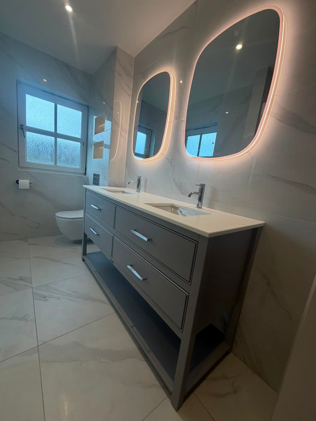 Marble tiled bathroom with a twin vanity and backlit mirrors, fitted by Caro Bathrooms
