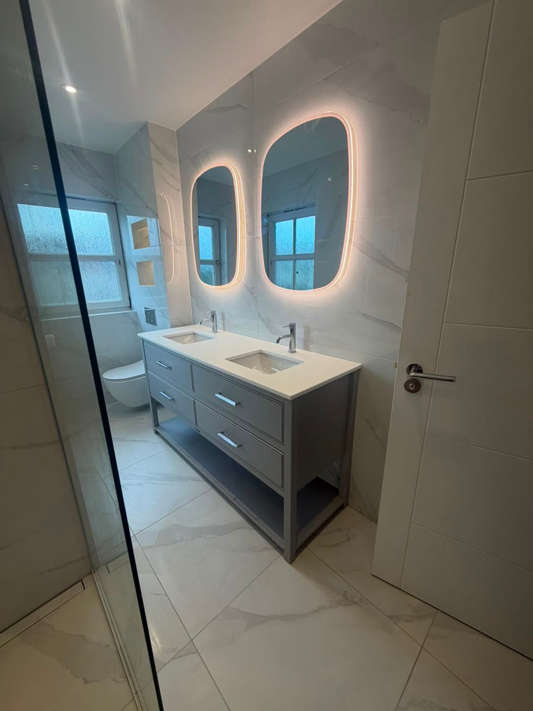Marble tiled bathroom with a twin vanity and backlit mirrors, fitted by Caro Bathrooms