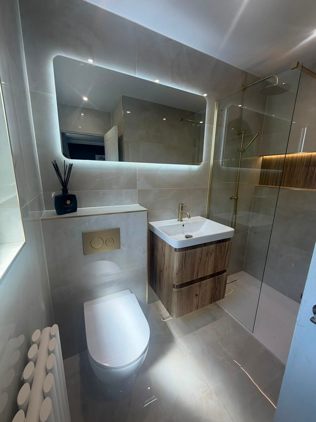 Grey tiled shower room with a wall-hung vanity and a backlit mirror, fitted by Caro Bathrooms