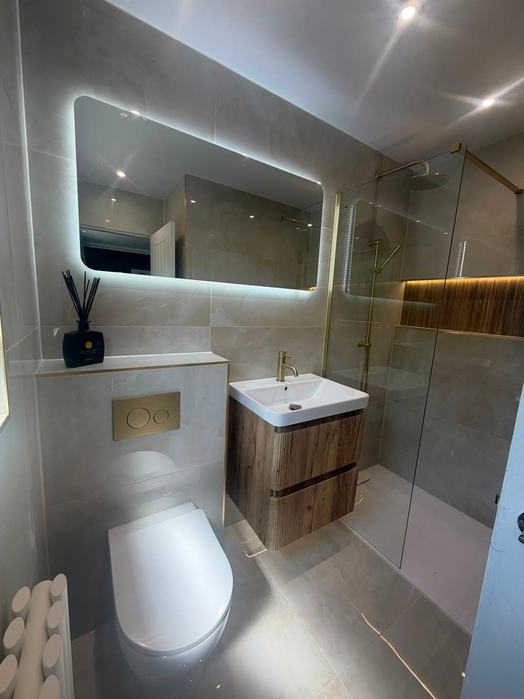 Grey tiled shower room with a wall-hung vanity and a backlit mirror, fitted by Caro Bathrooms